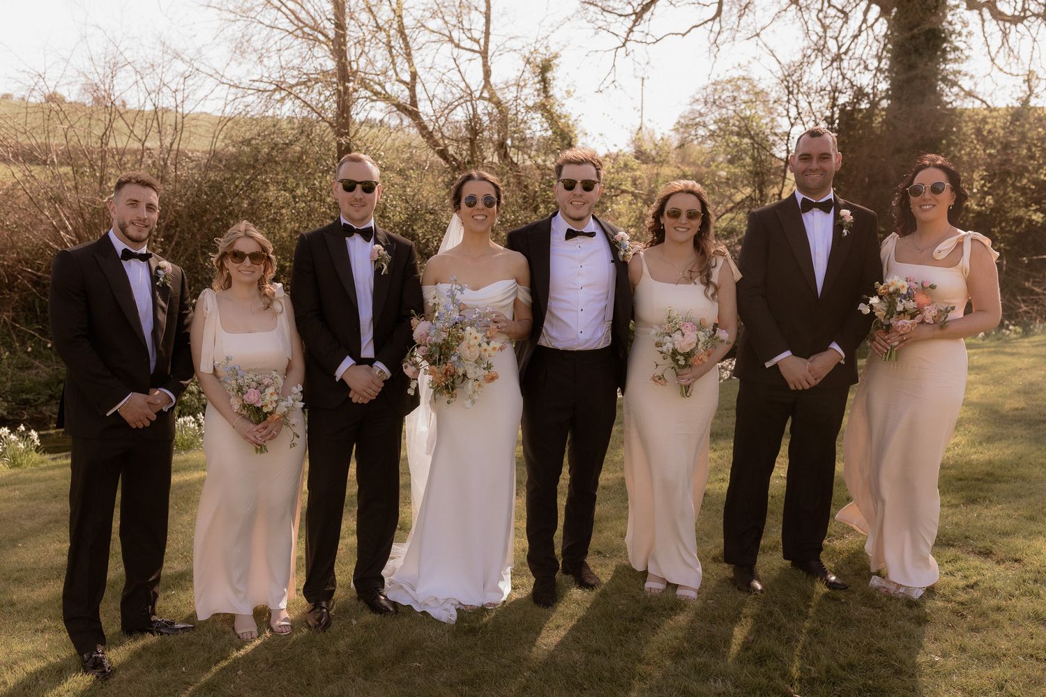 A wedding party poses outdoors in formal attire with bridesmaids in cream dresses.