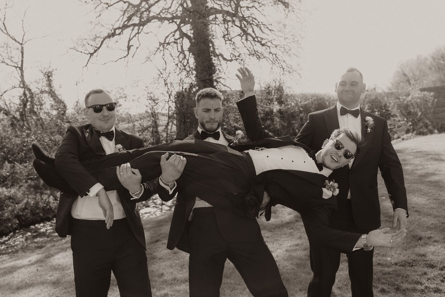 Groomsmen in black tuxedos pose playfully for a black and white photo outdoors.