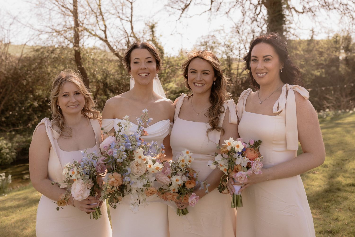 Four bridesmaids in cream-colored dresses hold pastel bouquets while smiling outdoors.