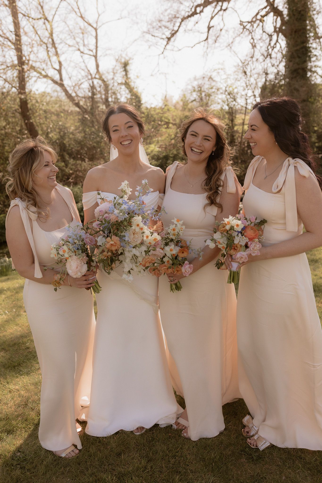 A group of bridesmaids in cream-colored dresses hold pastel bouquets while laughing together outdoors.