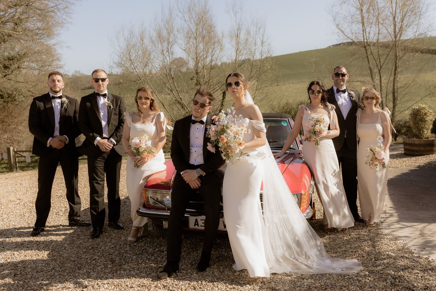Wedding party poses together in front of vintage cars on a countryside driveway.