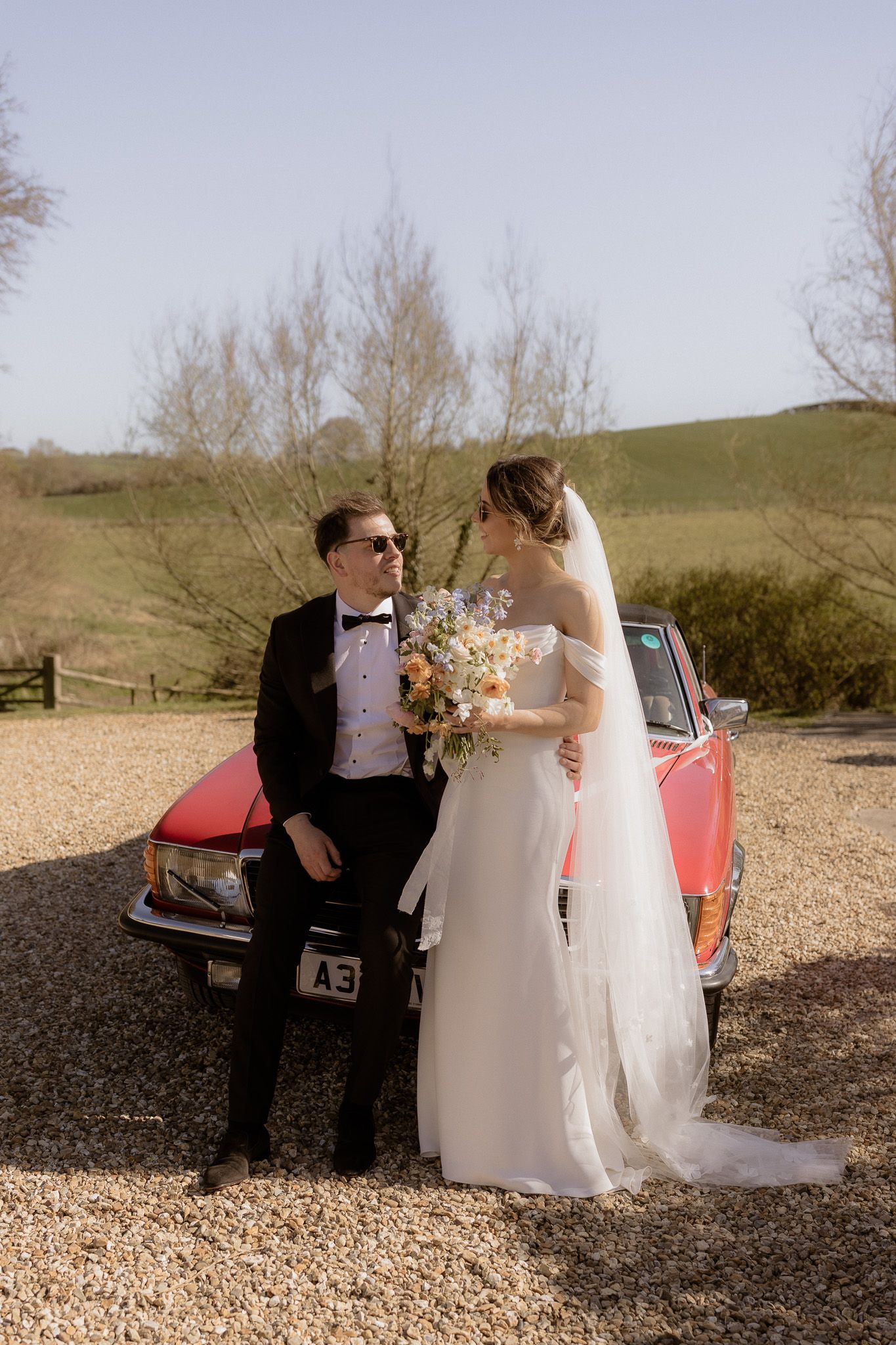 A wedding couple poses with a vintage red car against a countryside landscape.