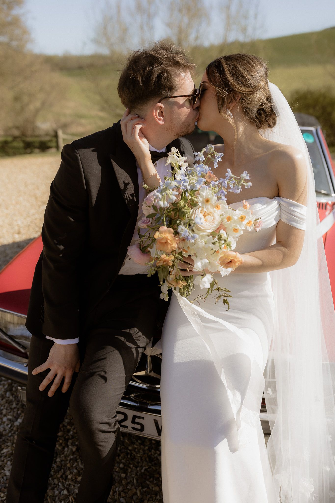 Romantic newlywed moment captured by vintage red car with spring flowers and countryside backdrop.