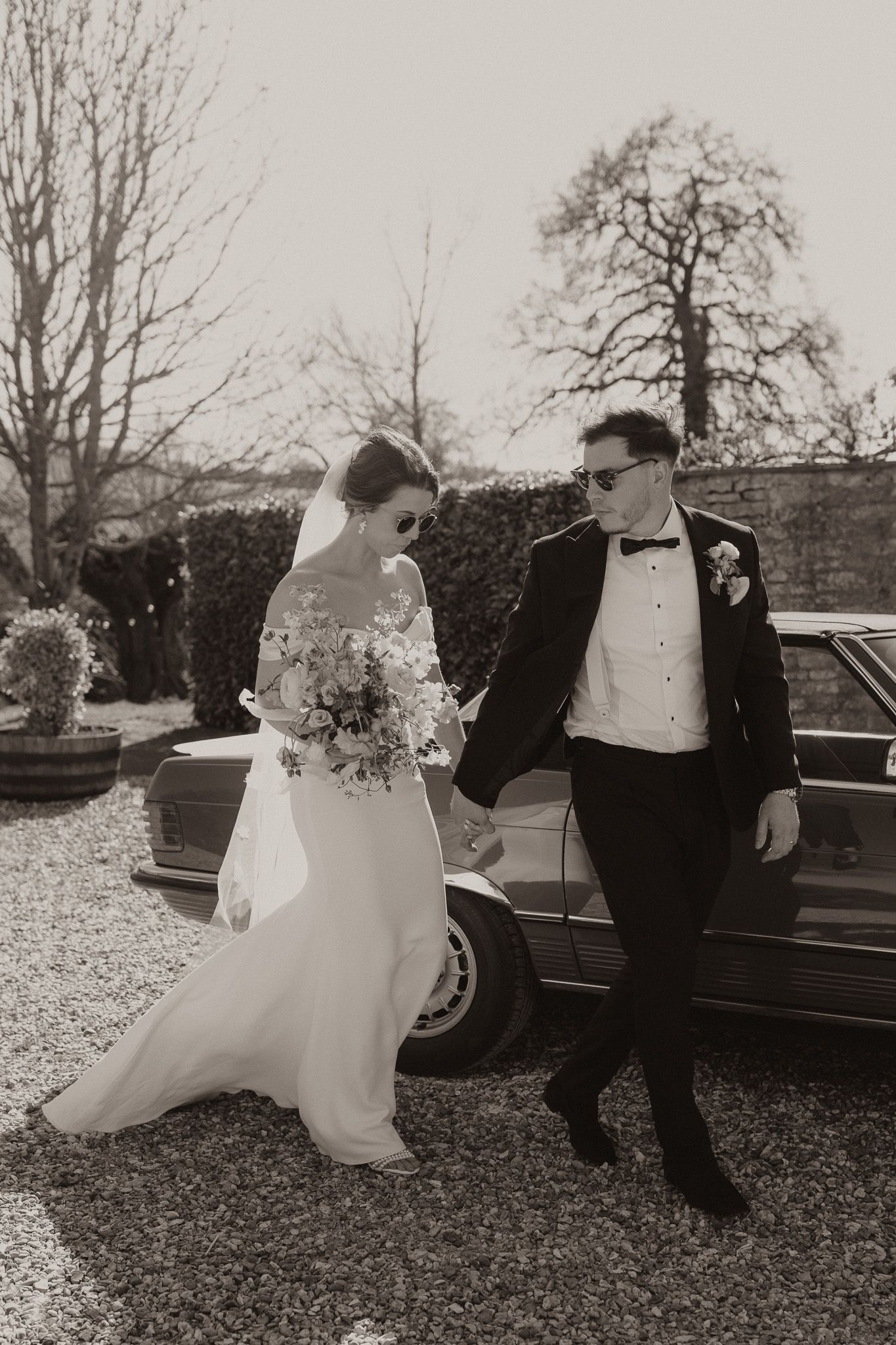 A black and white portrait of newlyweds standing beside a vintage car.