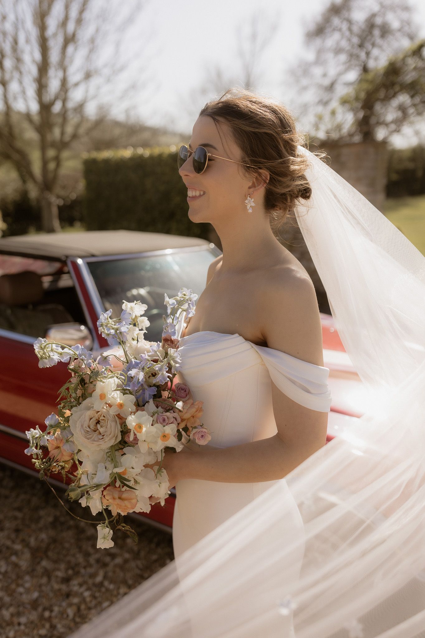 Bride in off-shoulder gown holding bouquet beside vintage car on sunny day.