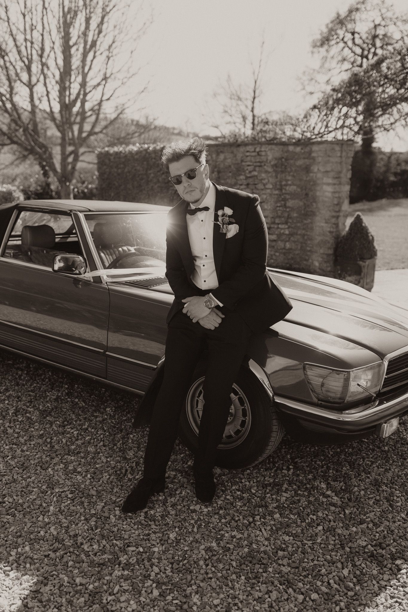 Classic black and white photo of groom in tuxedo leaning against vintage Mercedes car.