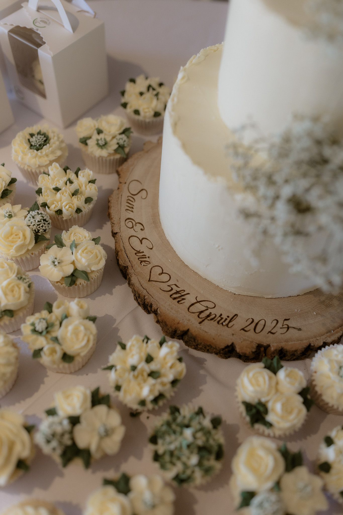 White wedding cake on a wooden slice surrounded by delicate cream rose decorations and floral arrangements.