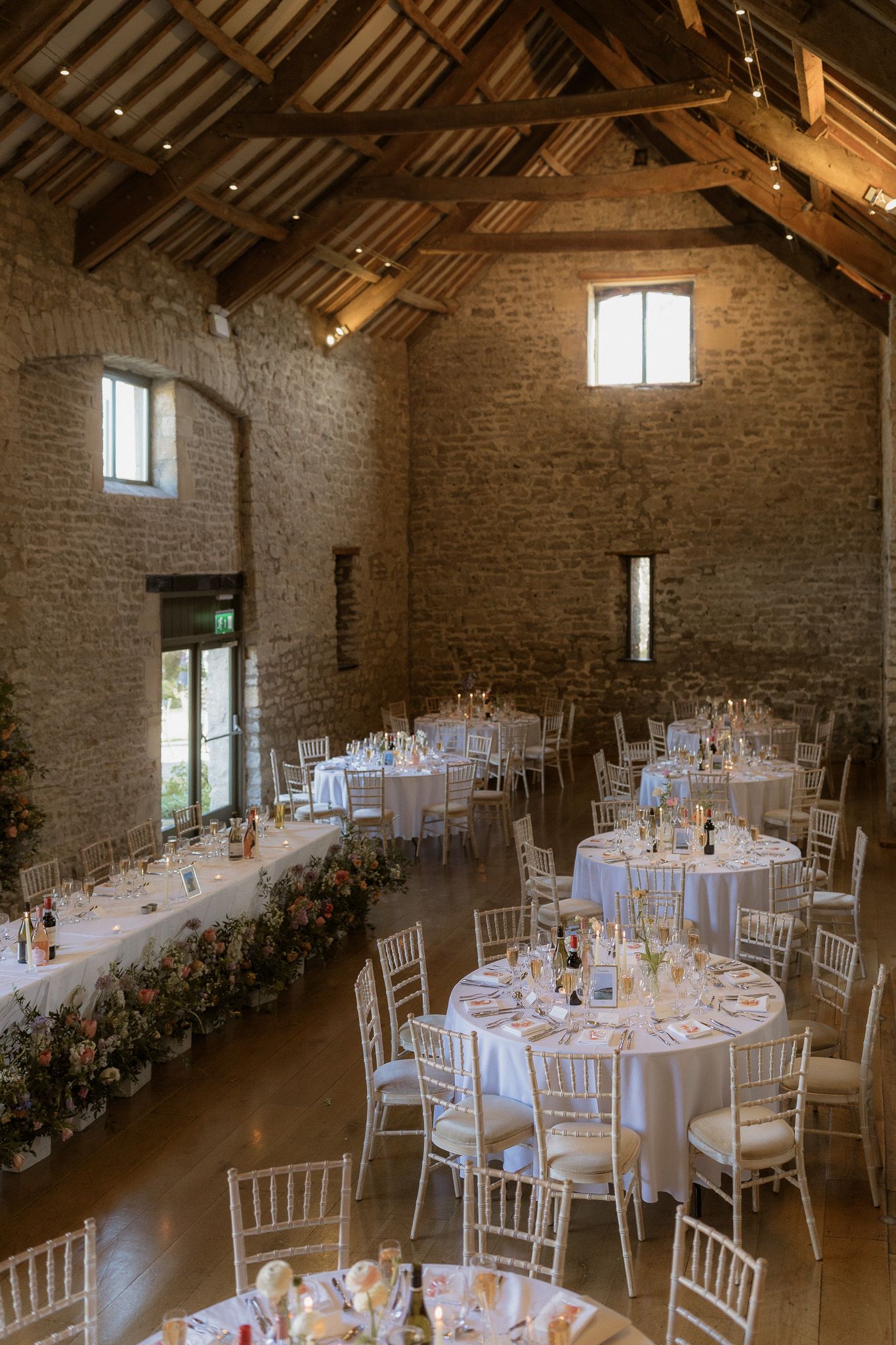 Rustic stone barn wedding reception setup with elegant round tables and white chairs under exposed wooden beams.