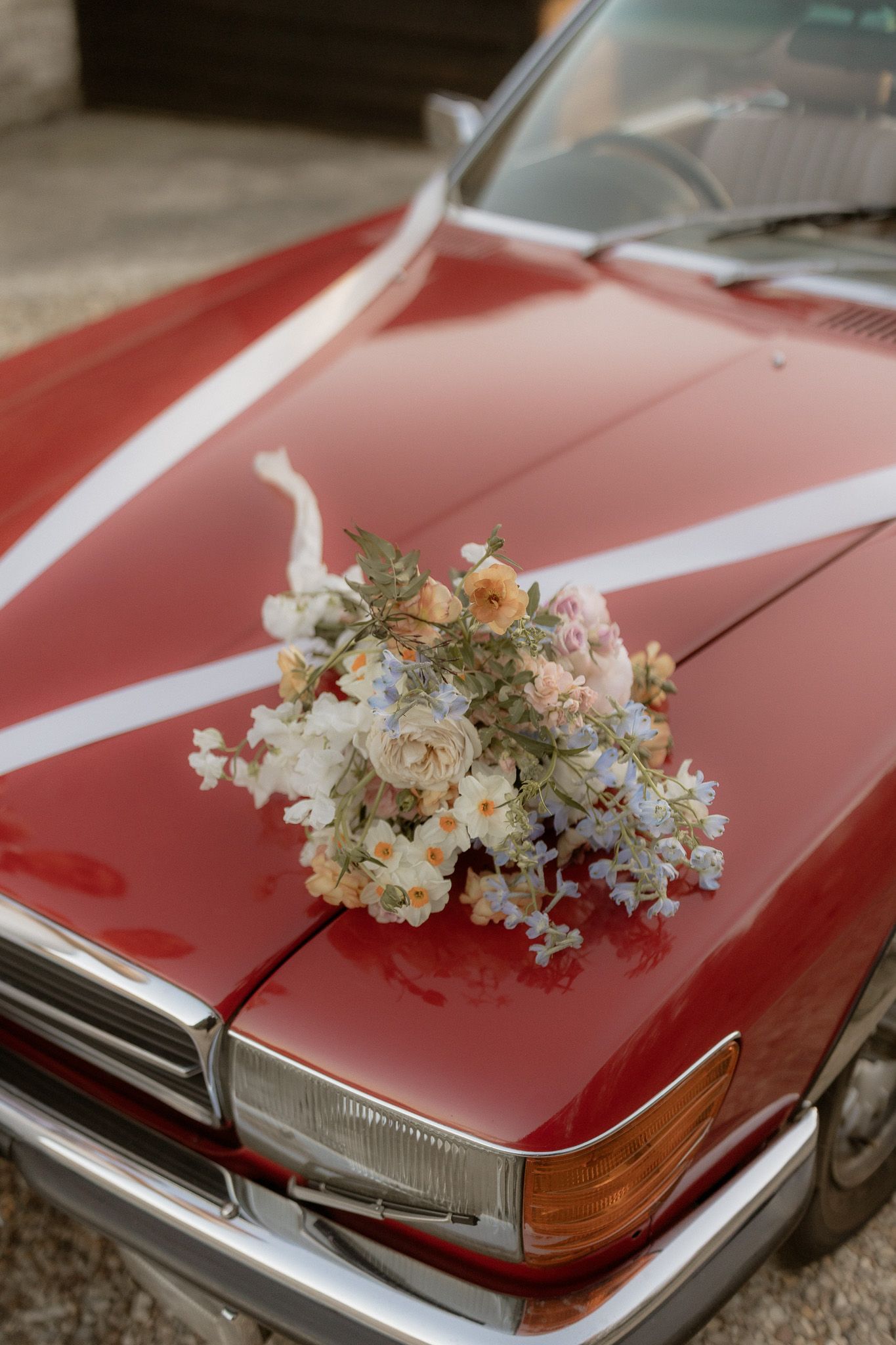 A delicate floral bouquet with white and peach blooms rests on the hood of a shiny red vintage car.