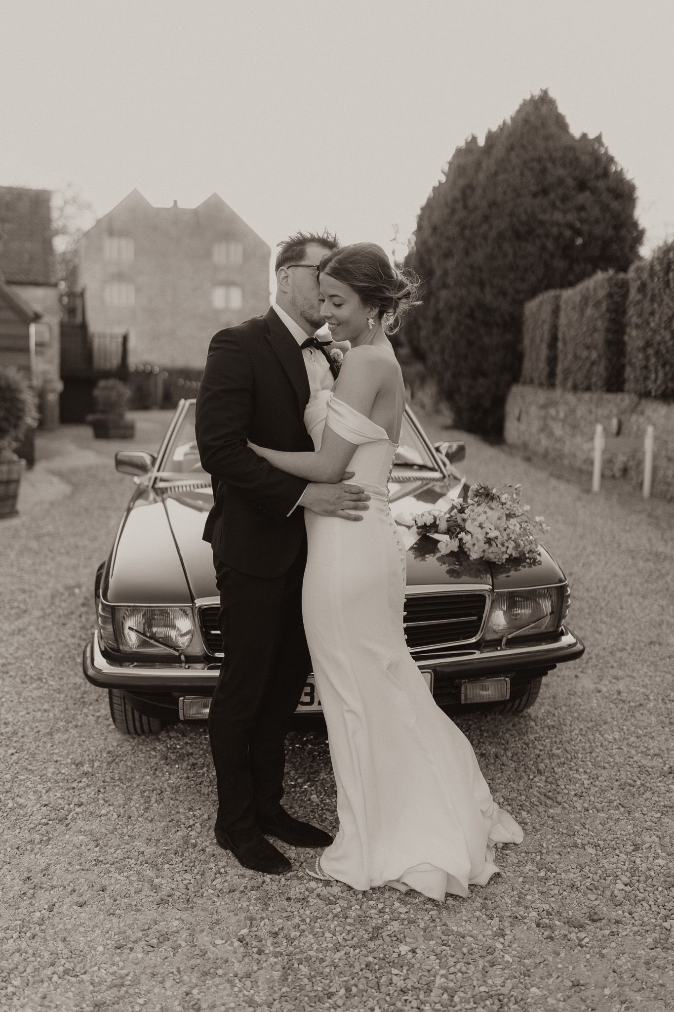 A romantic black and white wedding photo of a couple embracing by a classic car.