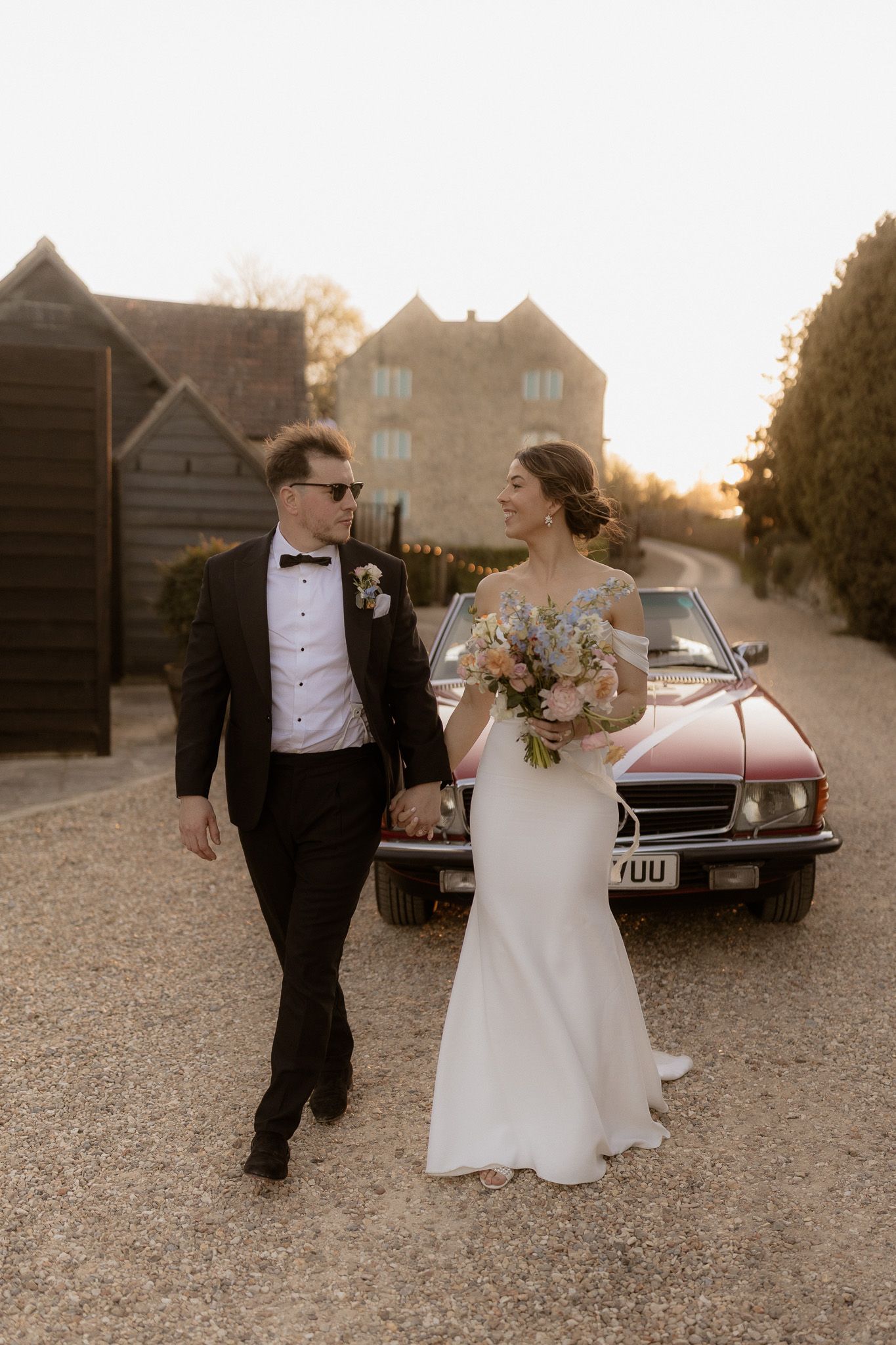 A couple walks together on a gravel path with a vintage car and stone buildings in the background at sunset.