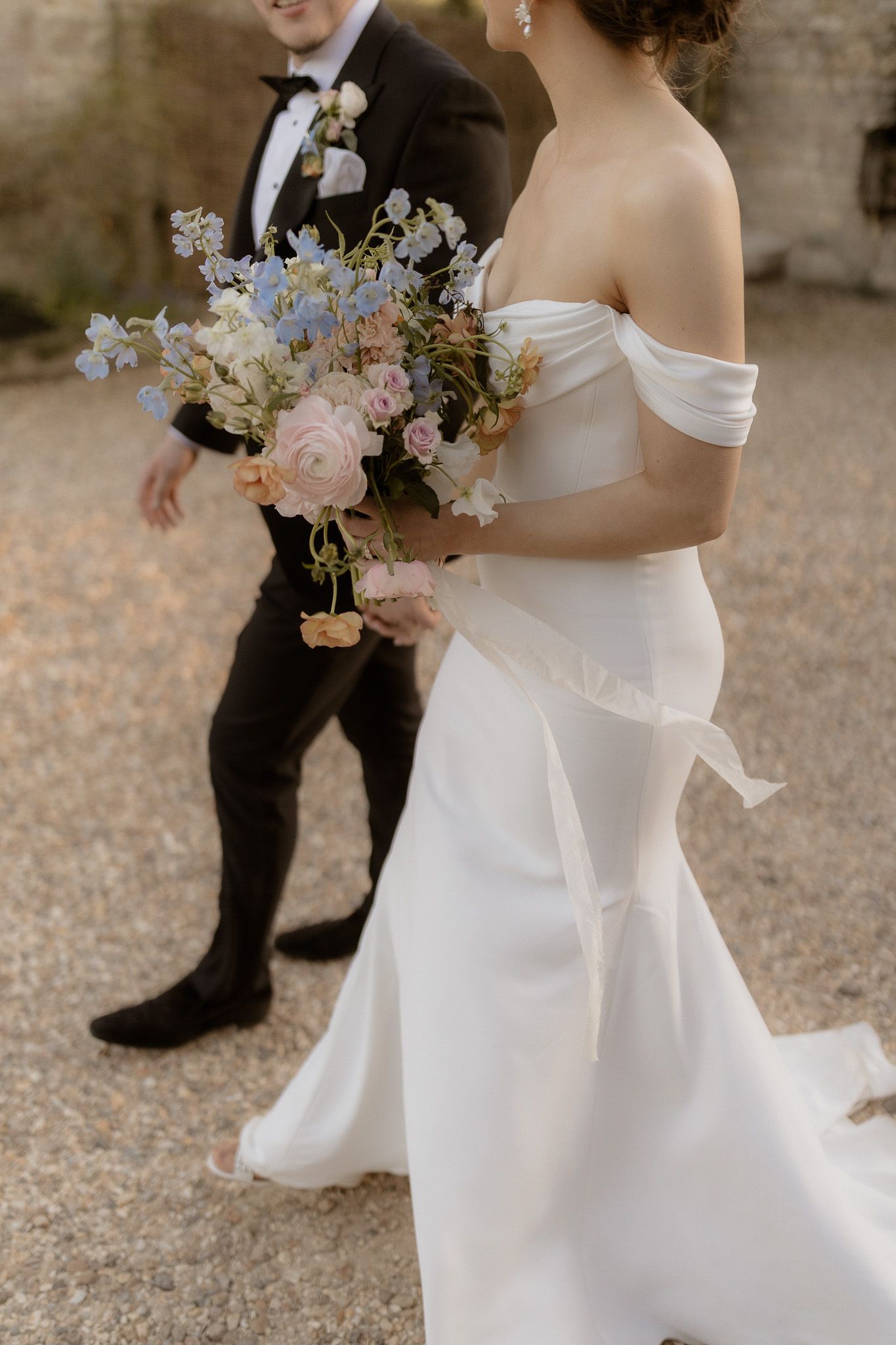 Elegant bride in off-shoulder white gown holding pastel floral bouquet next to groom in black tuxedo.