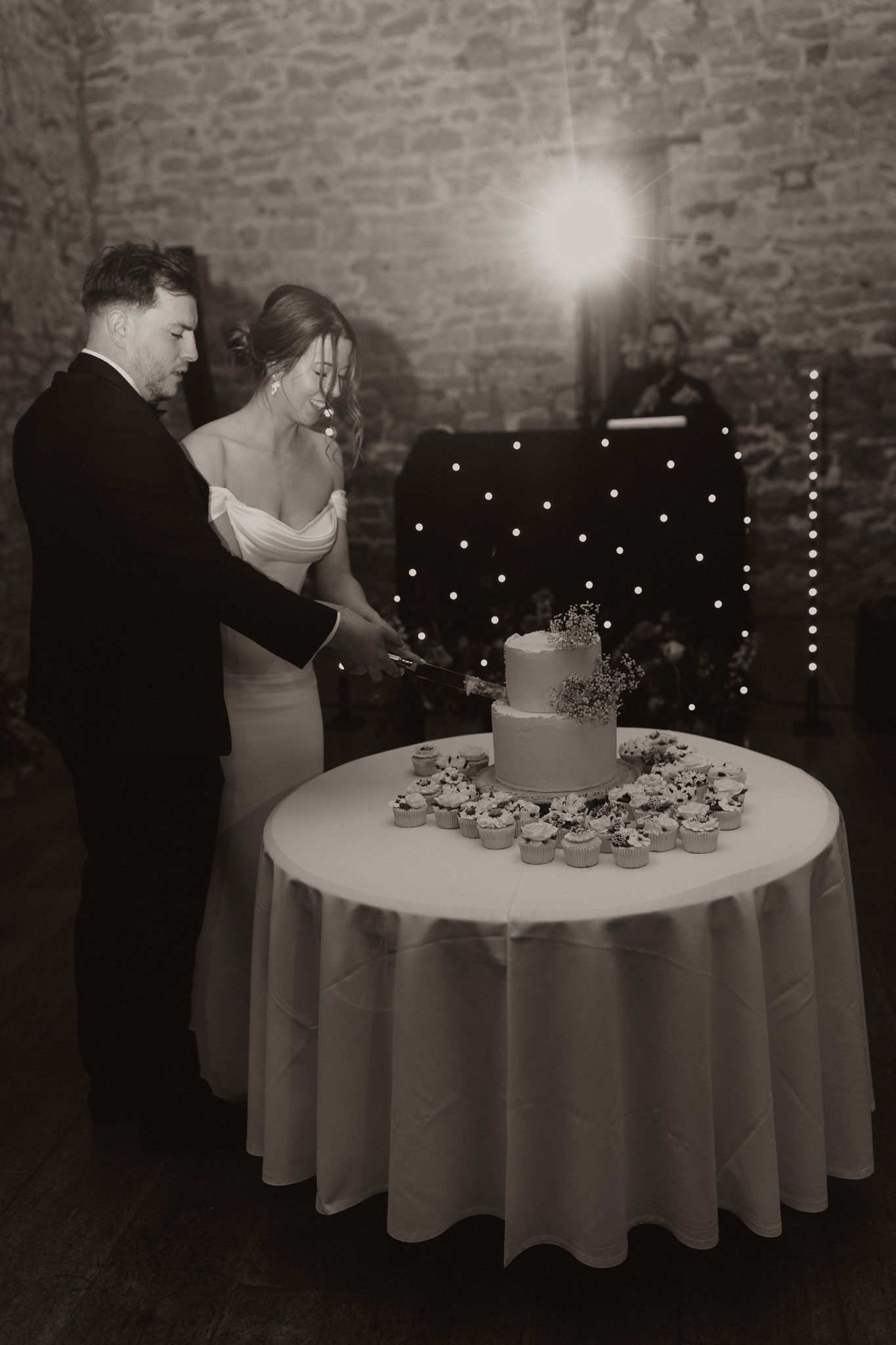 Black and white photo of wedding cake cutting ceremony in an intimate brick-walled venue.