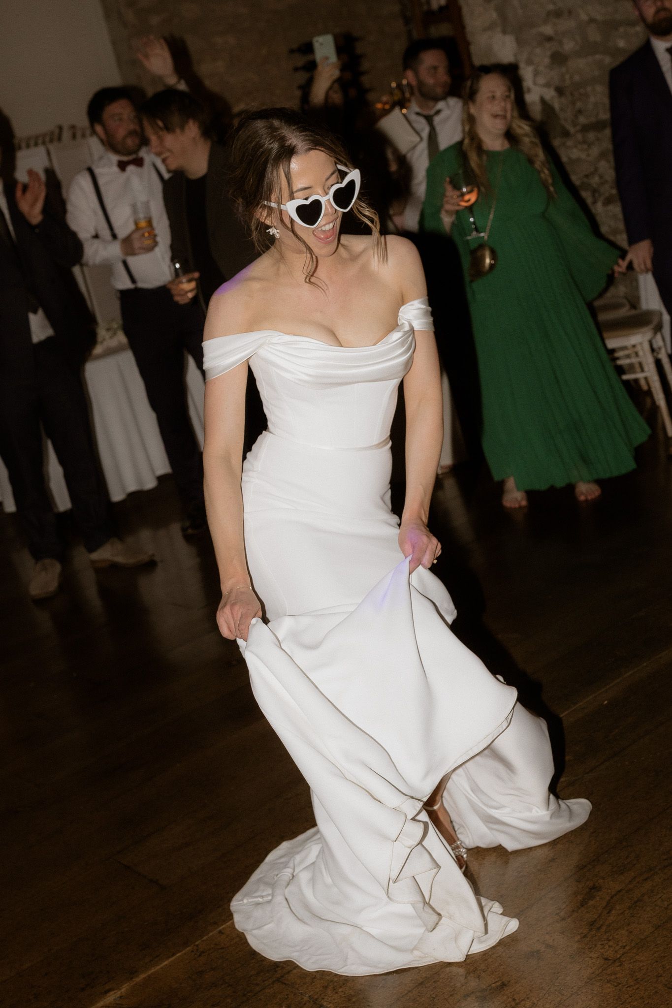 Dancing bride wearing sunglasses and white off-shoulder wedding dress at reception party.