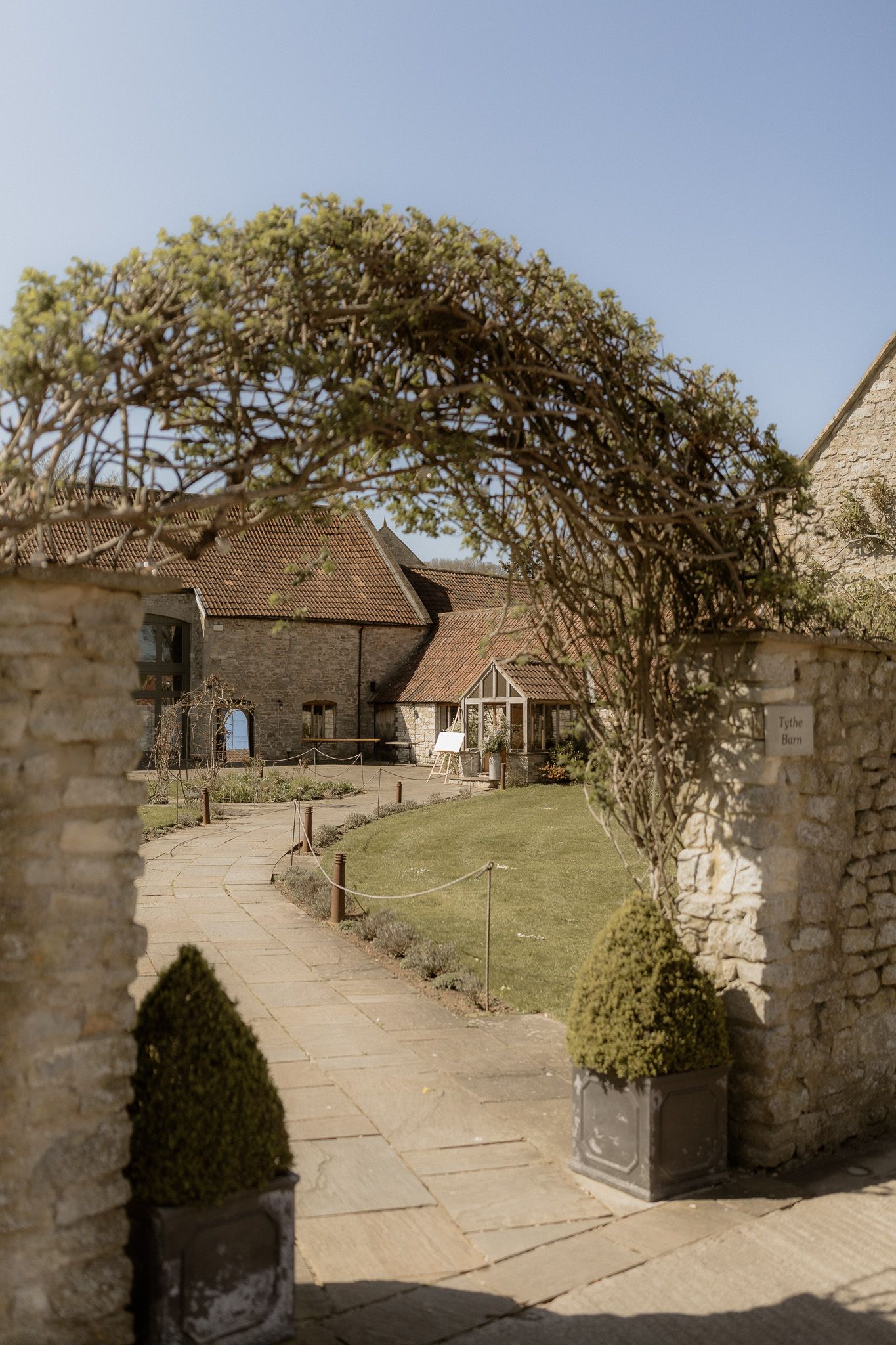 Stone archway covered in climbing vines leads to a winding path through manicured garden grounds.
