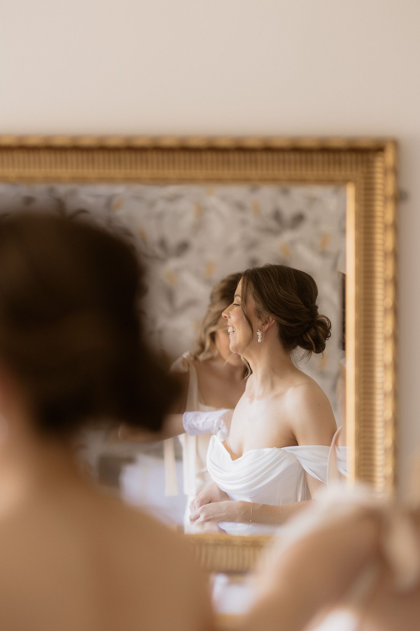 A reflection in an ornate gold mirror shows a bride in an elegant off-shoulder white dress with her hair in a romantic updo.