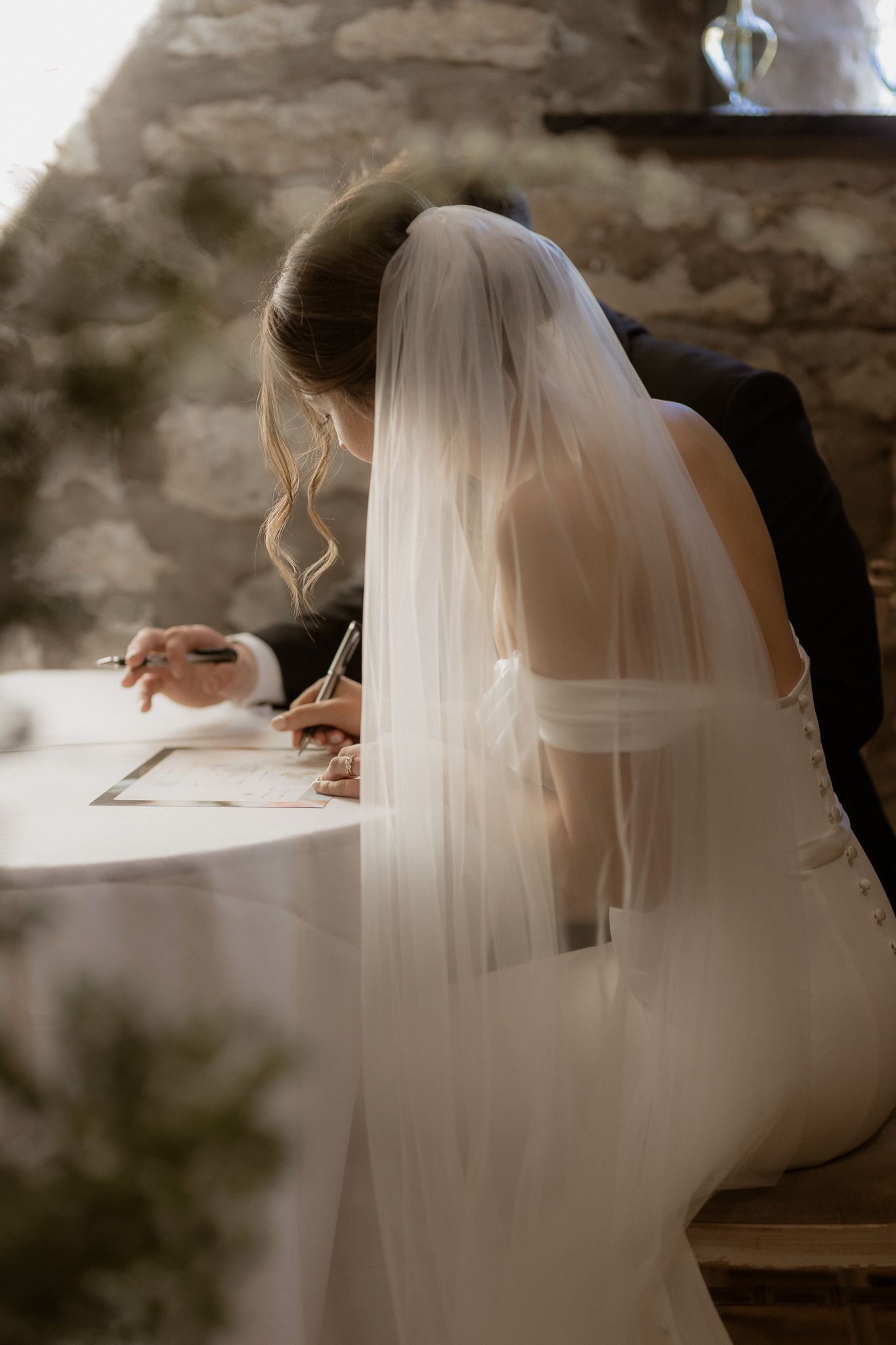 Bride in white dress and veil signing wedding documents at rustic stone venue.