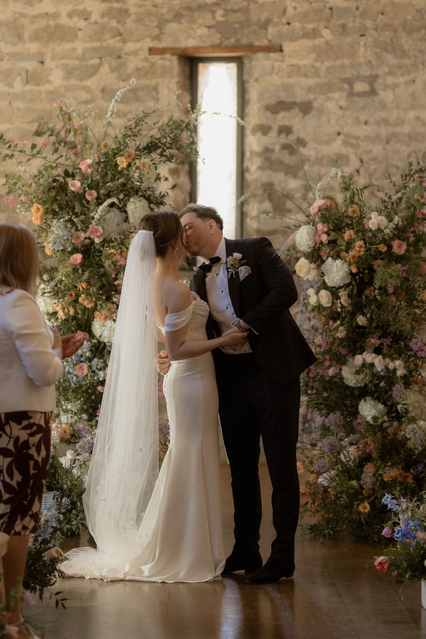 A wedding ceremony moment under a floral arch with peach and white blooms in a stone venue.
