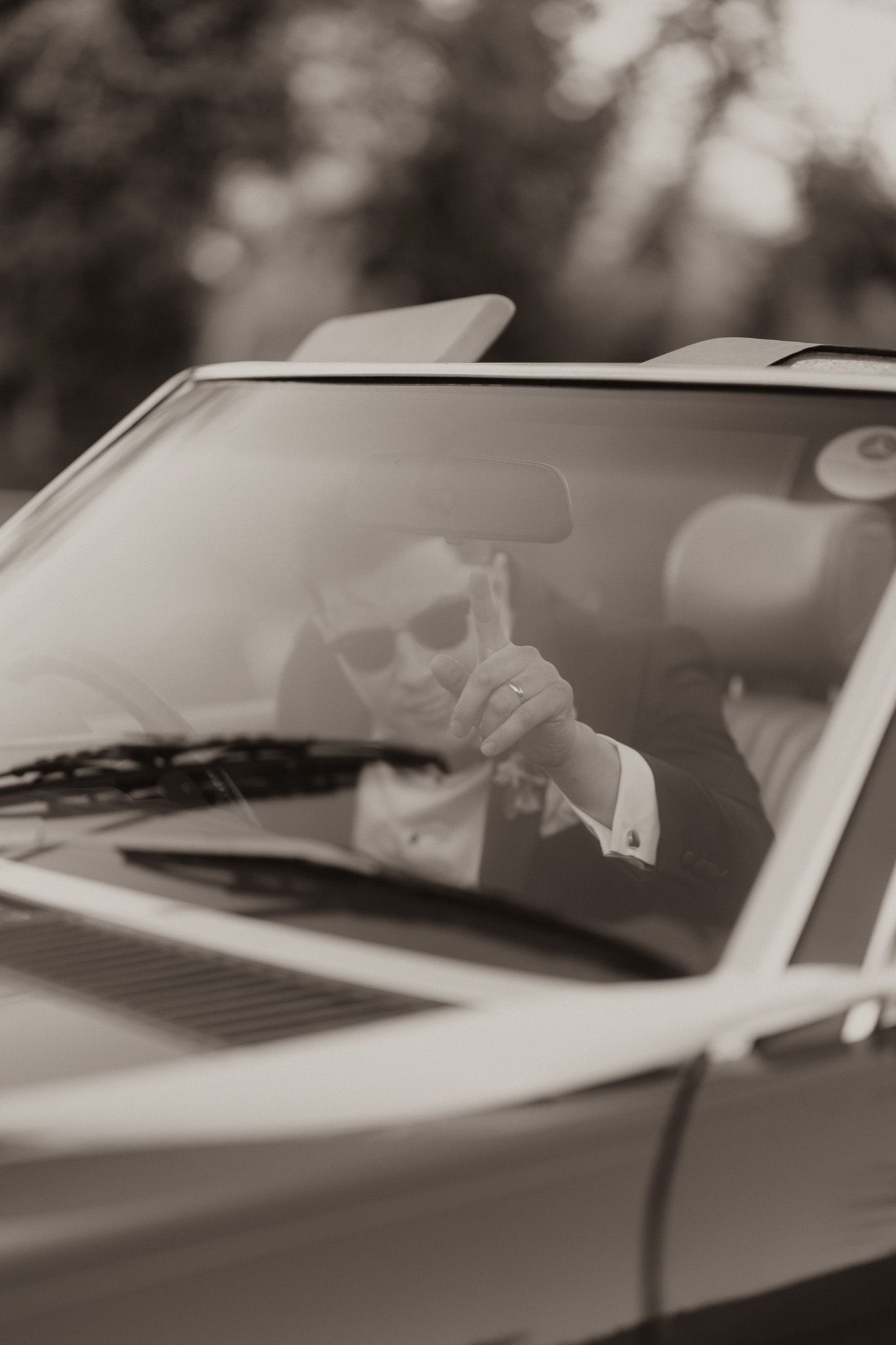 Person wearing sunglasses gestures from inside a luxurious car in a black and white photograph.