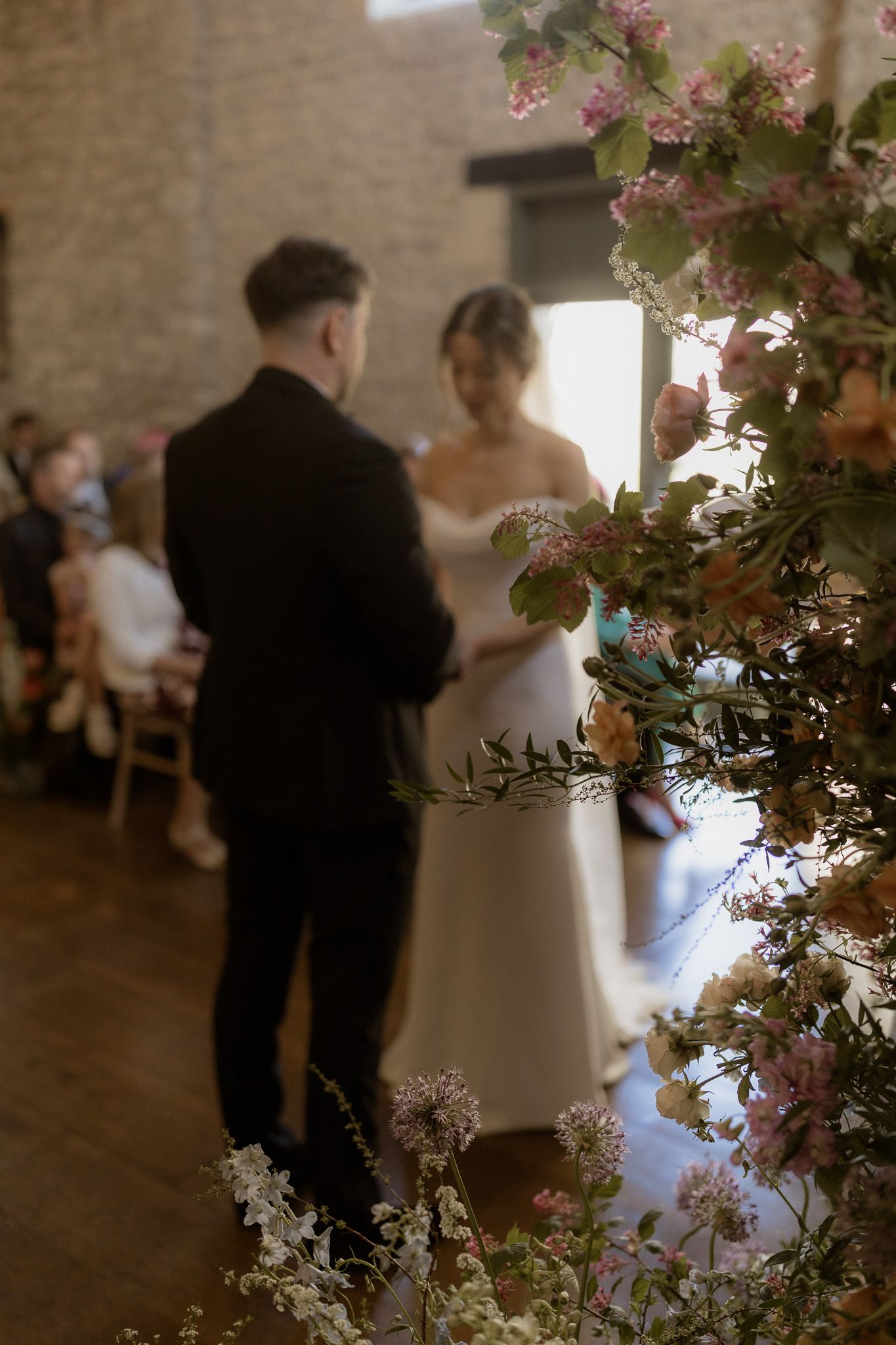 A couple in wedding attire embraces during a ceremony framed by delicate floral arrangements in a rustic brick venue.