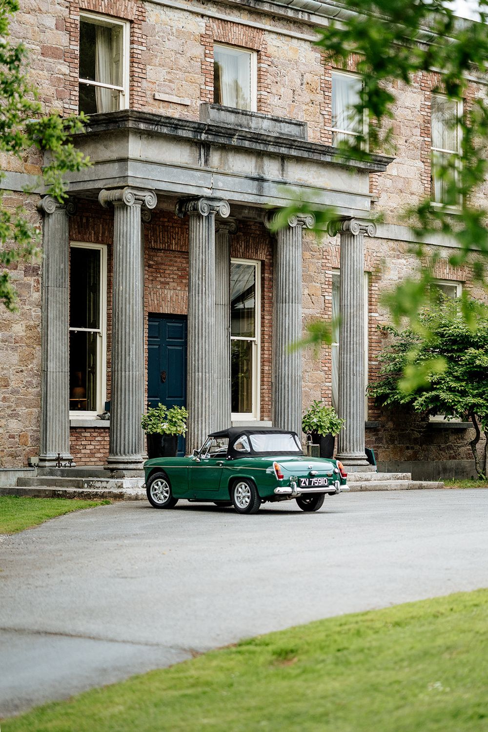 A vintage green sports car parked in front of a historic stone building with classical columns and red brick facade.