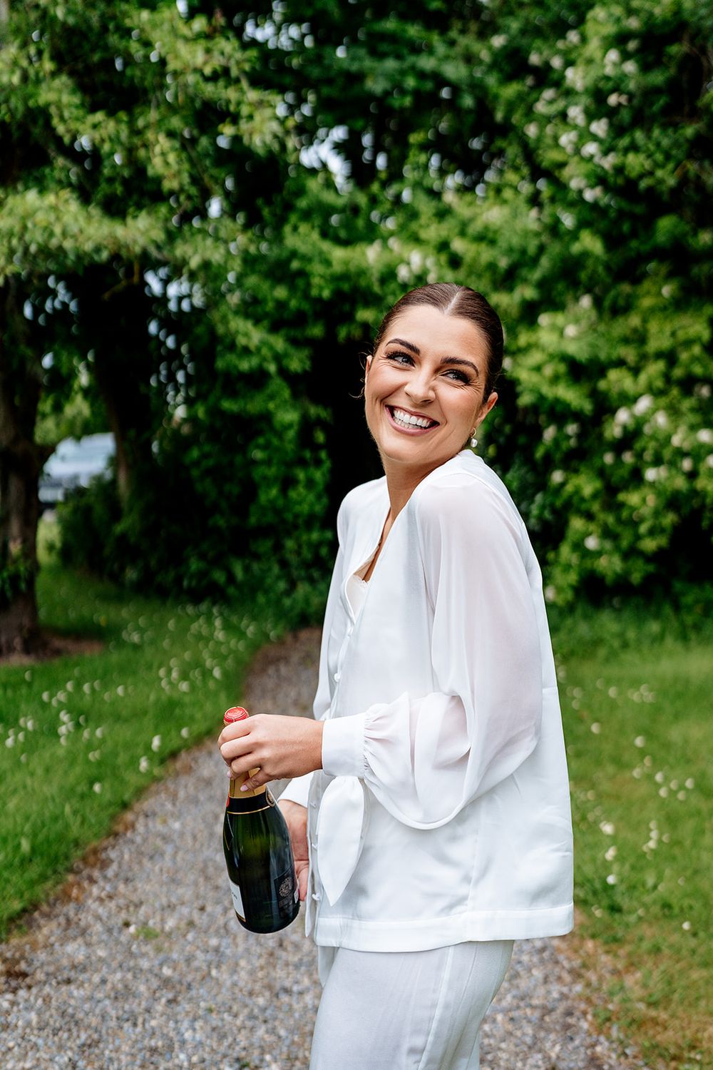 Person wearing a white outfit laughs joyfully while standing on a gravel path surrounded by lush green trees outdoors.