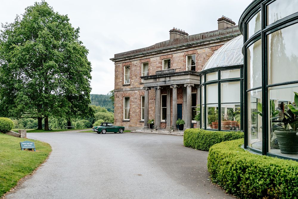 Elegant Victorian mansion with stone facade and curved glass conservatory extension set in manicured garden grounds.