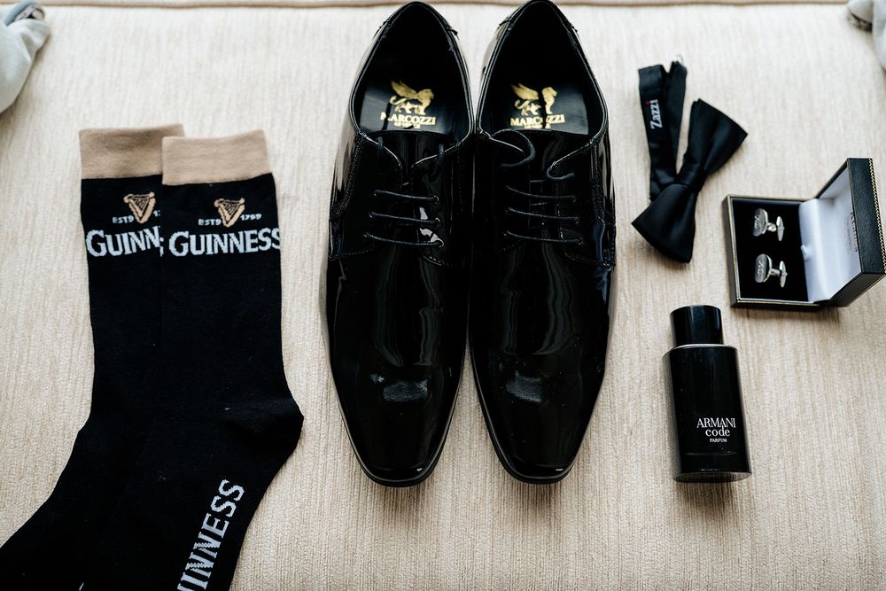Black formal dress shoes arranged with matching socks, cufflinks, and cologne on a light beige fabric surface.