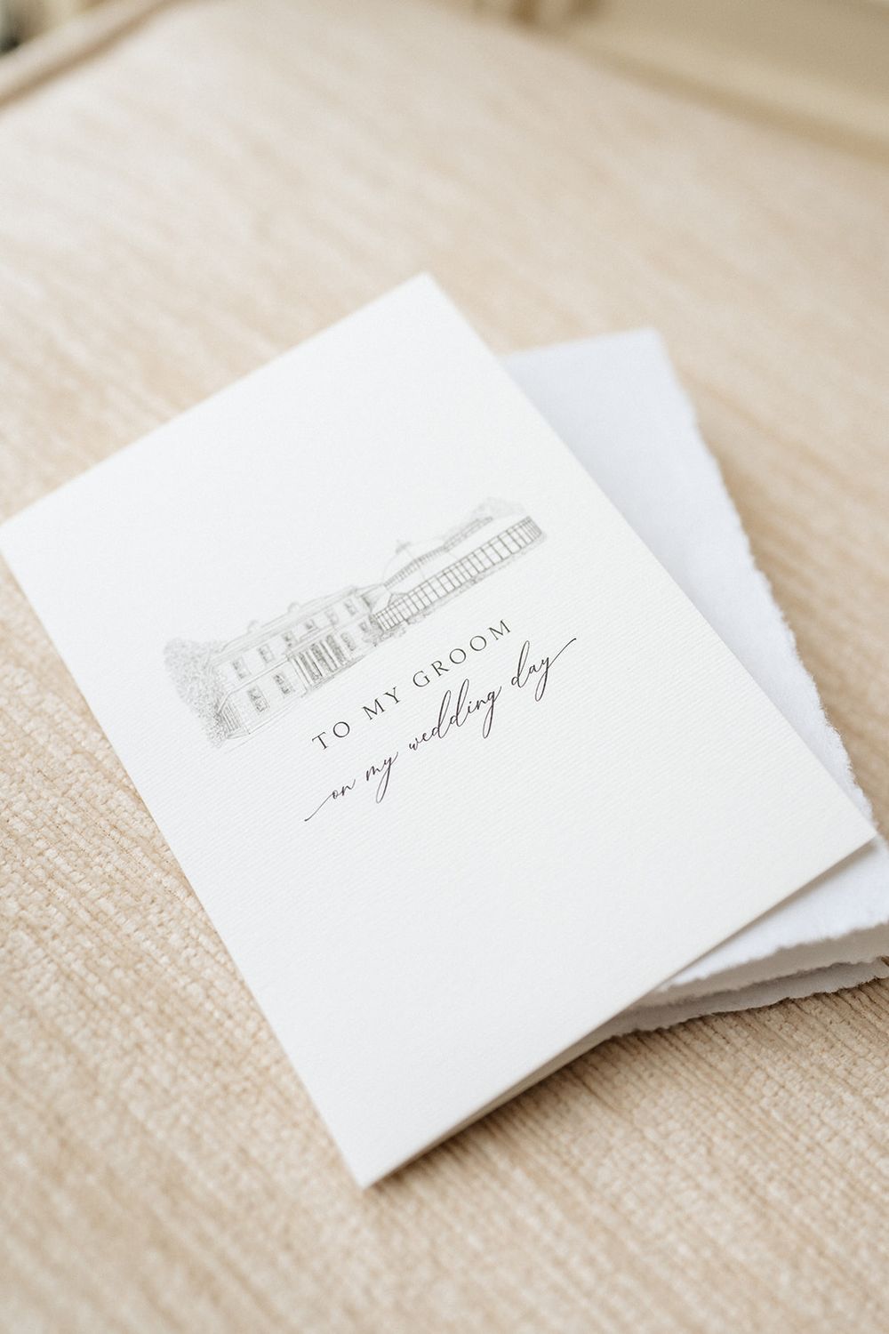 A minimalist white wedding card with elegant gray text and architectural sketch lies on a beige linen surface.