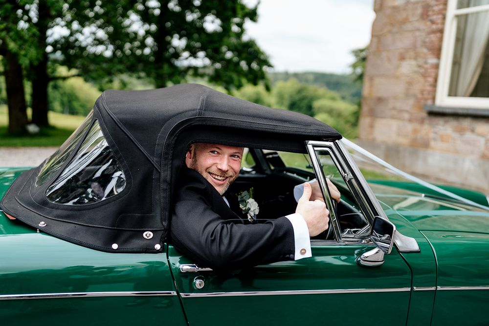 A person gives thumbs up while sitting in a classic green convertible car with black soft top.