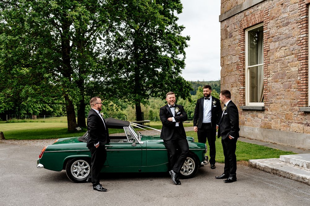 A group gathers around a classic green vintage car outside a brick building.
