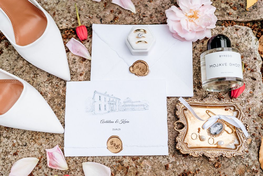 Wedding details flatlay with white shoes, invitation with custom venue illustration, wax seals and perfume bottle.