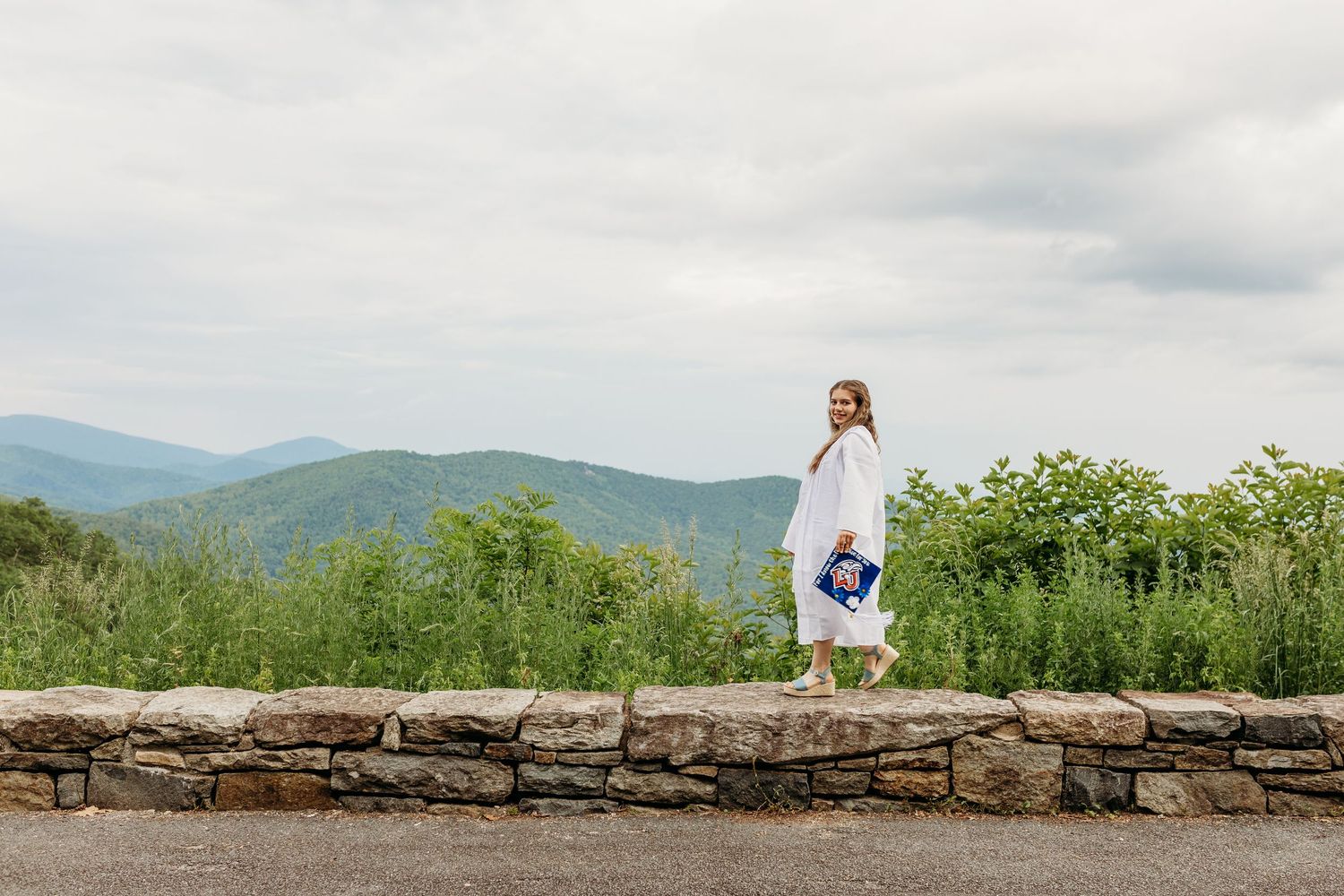 A woman in a white dress stands atop a stone wall with rolling mountains and lush greenery in the background.