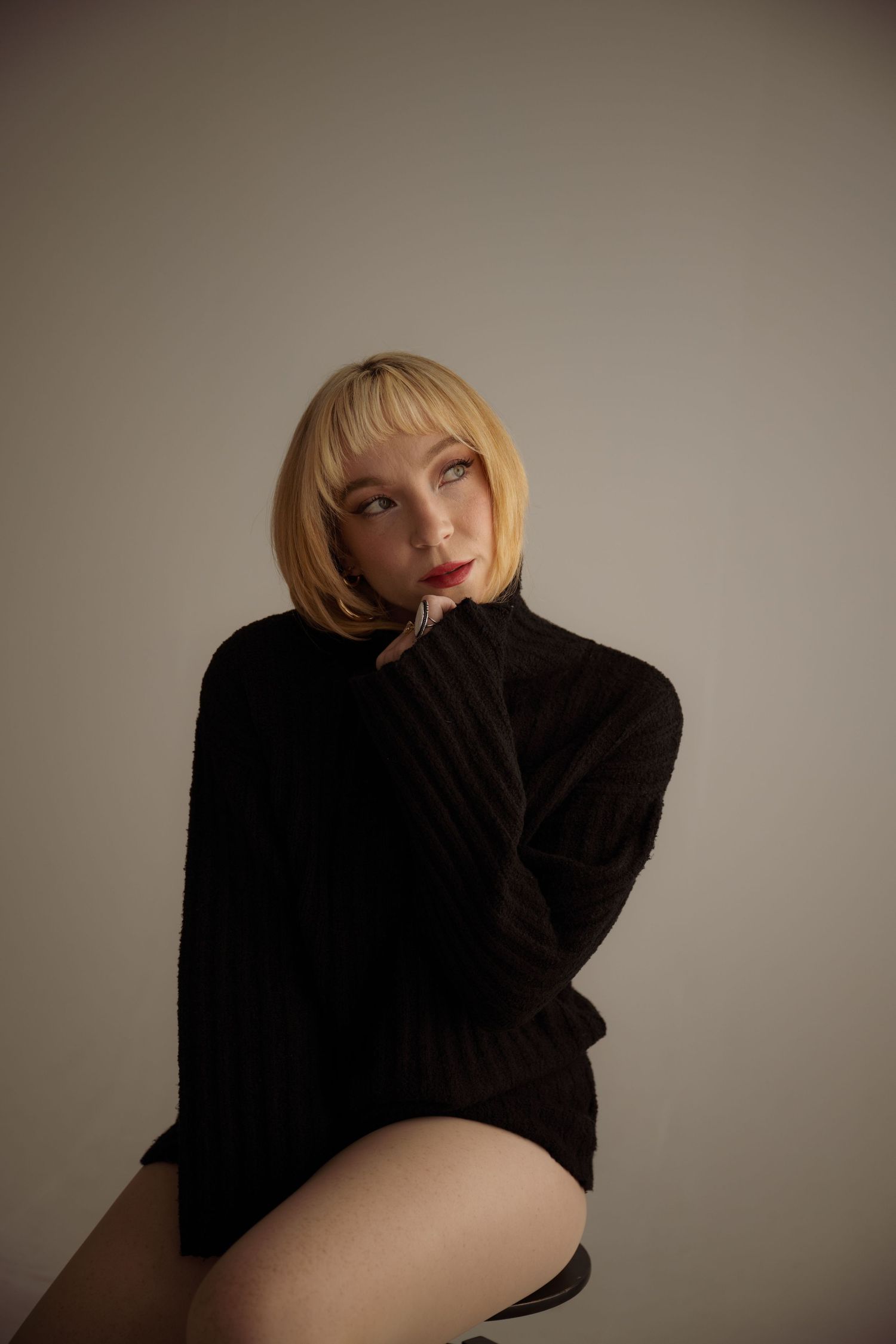 A model in dramatic lighting poses in a black sweater against a neutral background creating a moody portrait.