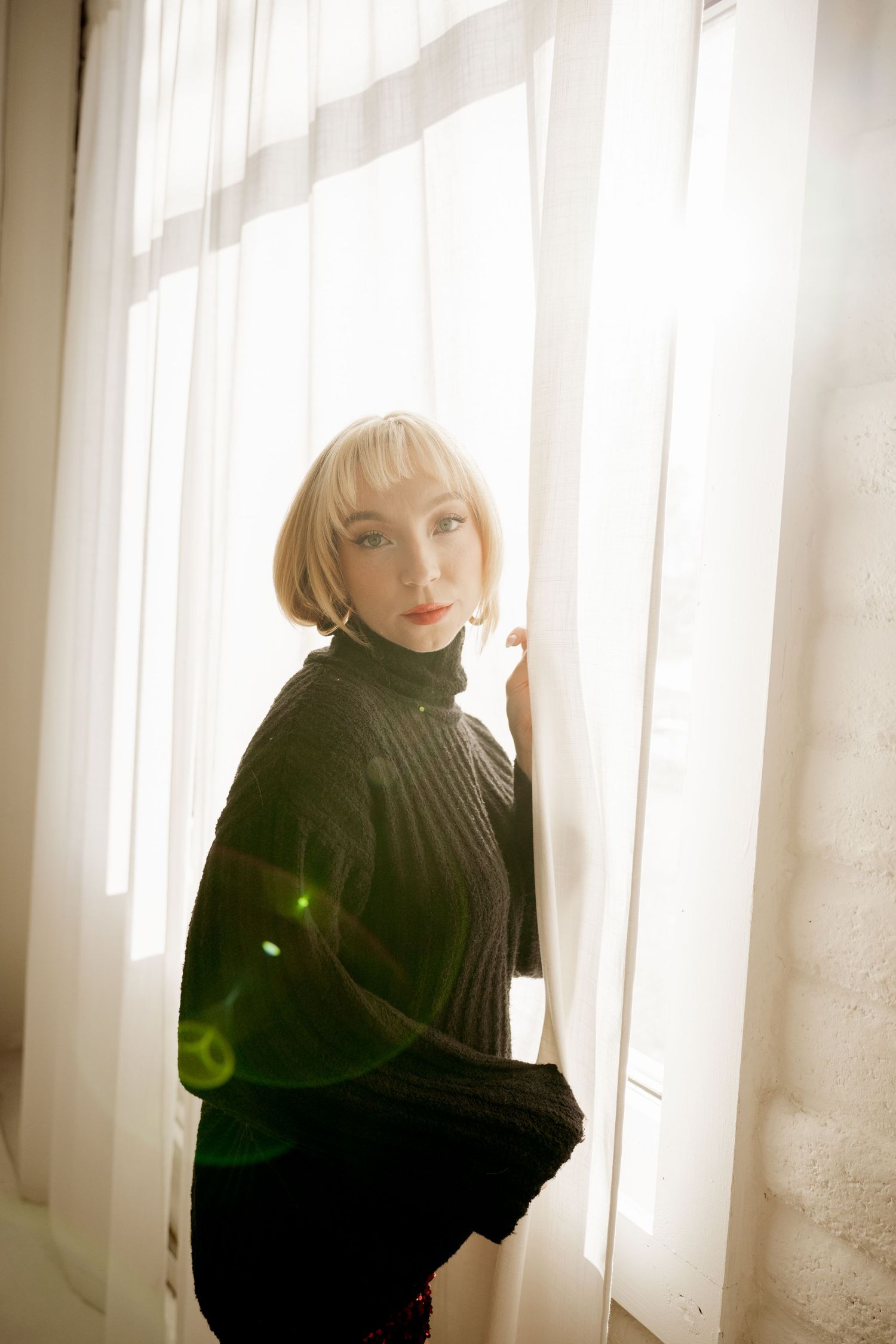 Figure in a black turtleneck sweater poses by a sunlit window with soft lighting and dreamy lens flare effects.