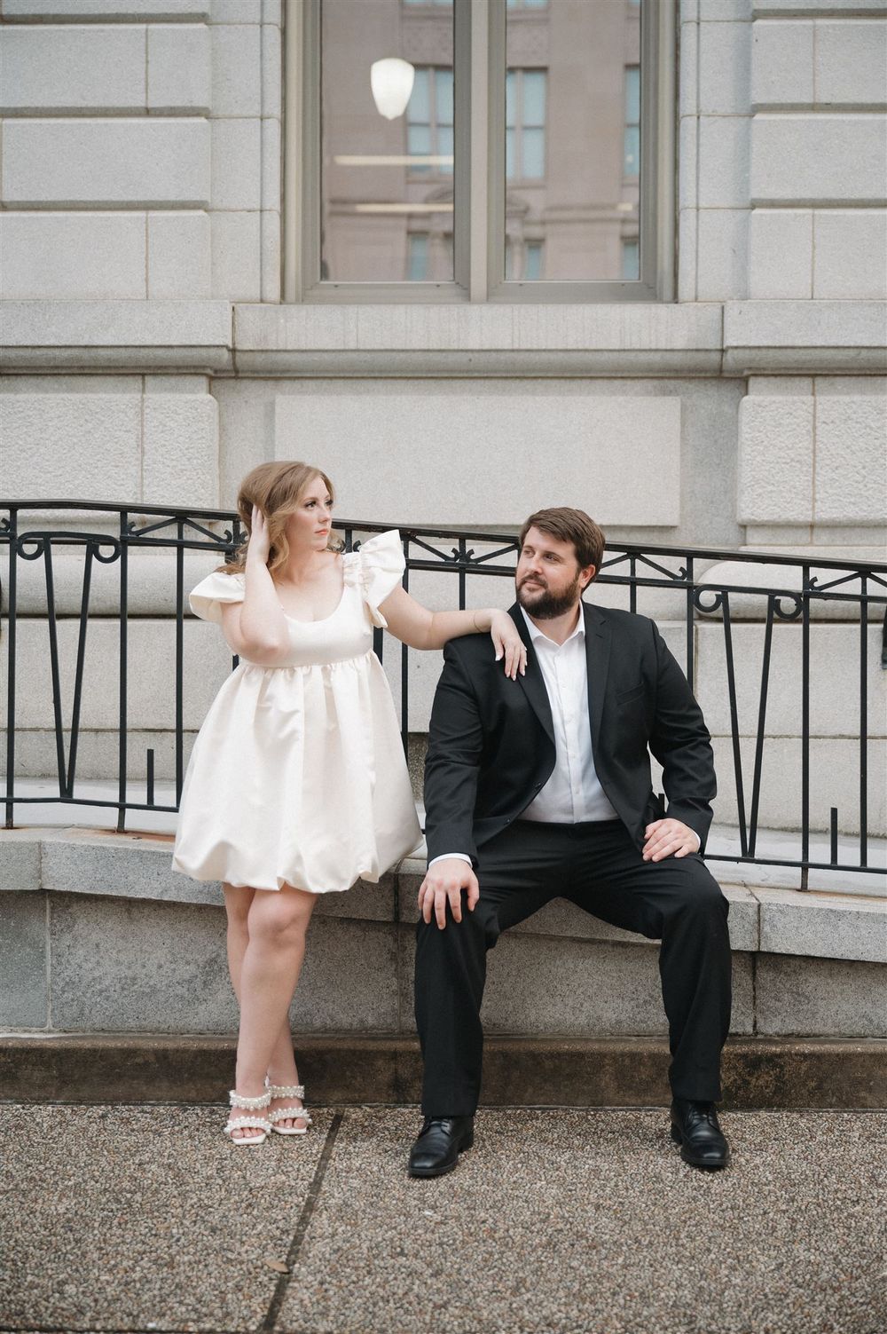 A couple poses together on stone steps outside a classic building, with her in a white dress and him in a dark suit.