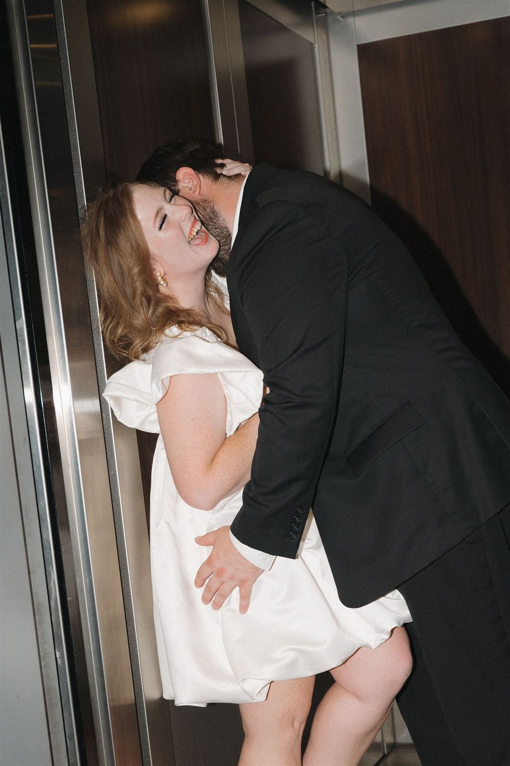A playful couple shares an intimate moment in an elevator, dressed in formal wear.