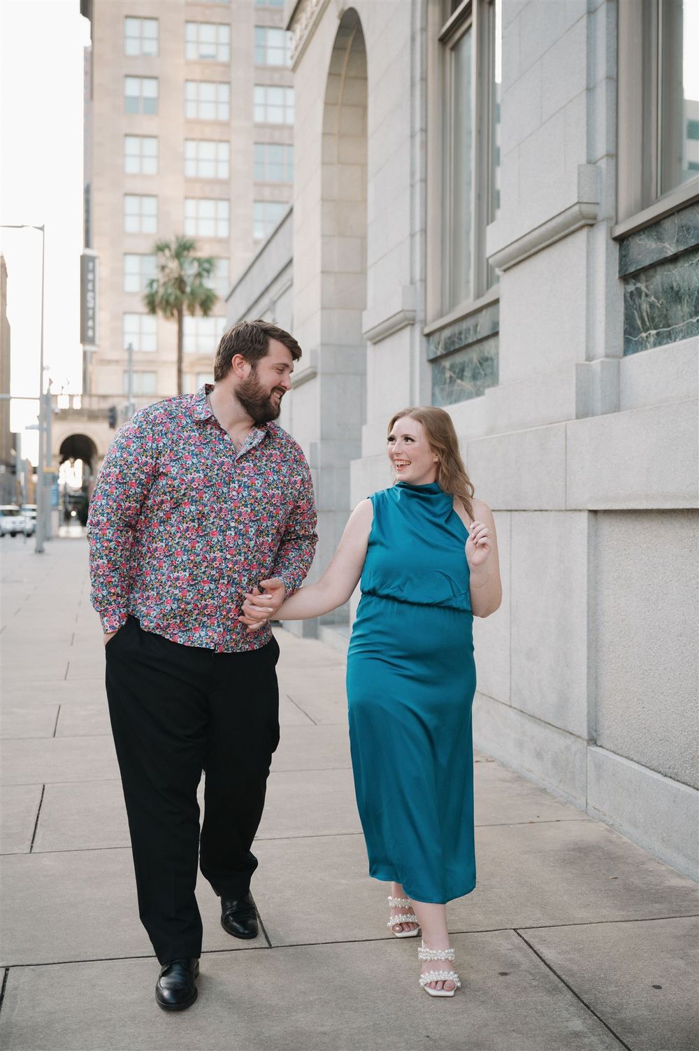 A couple walking and laughing together on a city sidewalk, with him in a floral shirt and her in a teal dress.
