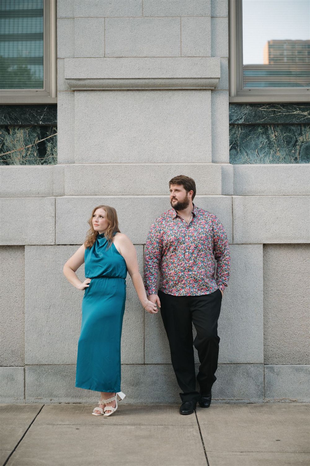 A stylish couple poses against a stone wall in formal attire, holding hands.