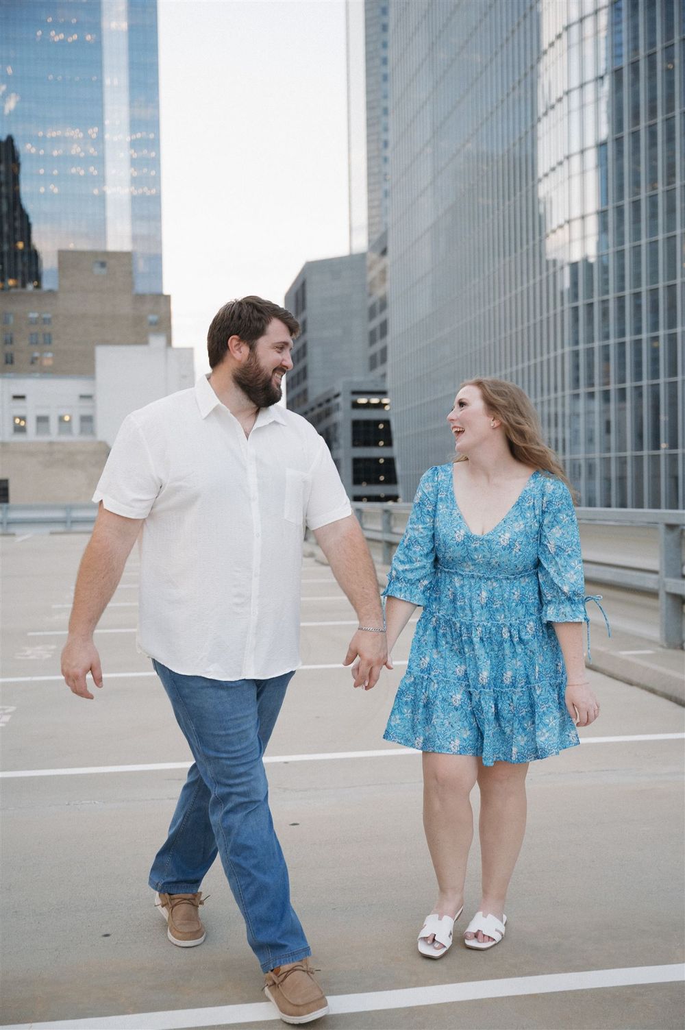 A couple walks hand in hand on a rooftop with modern city buildings in the background.