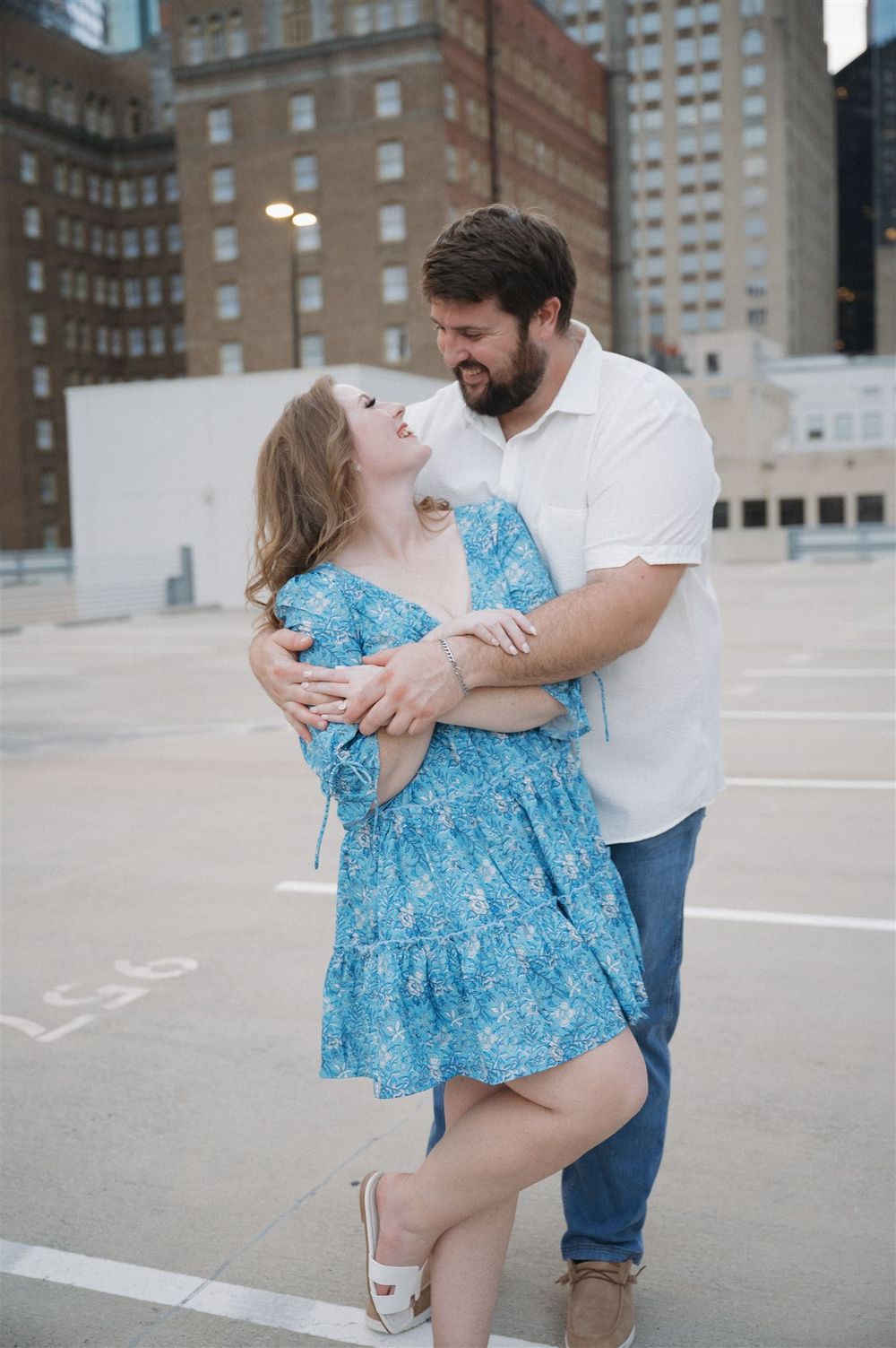A romantic couple embraces while dancing in a city parking lot, with the woman wearing a blue floral dress.