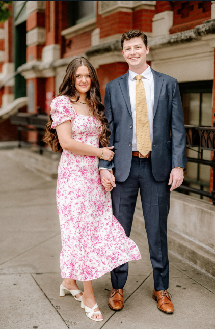 A couple stands in front of a historic brick building, with the woman in a pink floral dress and man in navy suit.
