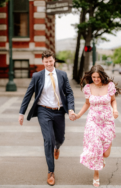 A couple in formal attire joyfully runs hand-in-hand down a city sidewalk on a summer day.