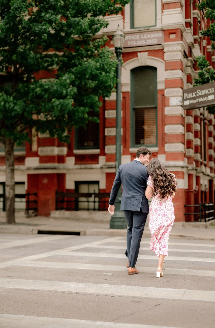 A couple walks away from a historic red brick building, with the woman wearing a pink floral dress and man in a suit.