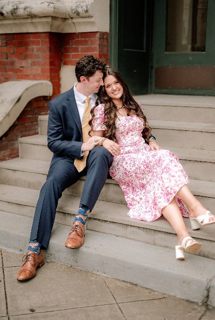 A couple sits intimately on concrete steps, with a man in a navy suit and a woman wearing a pink floral dress.