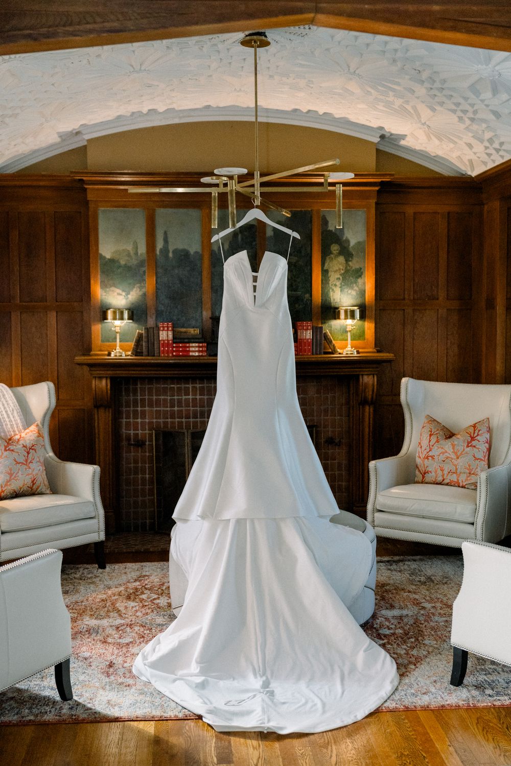 An elegant white wedding dress hangs between two gray armchairs in a wood-paneled room with decorative wallpaper details.