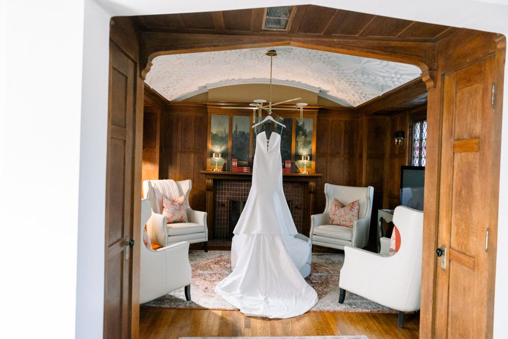 A white wedding dress hangs elegantly from a wooden beam in a rustic room with upholstered chairs and butterfly decor.