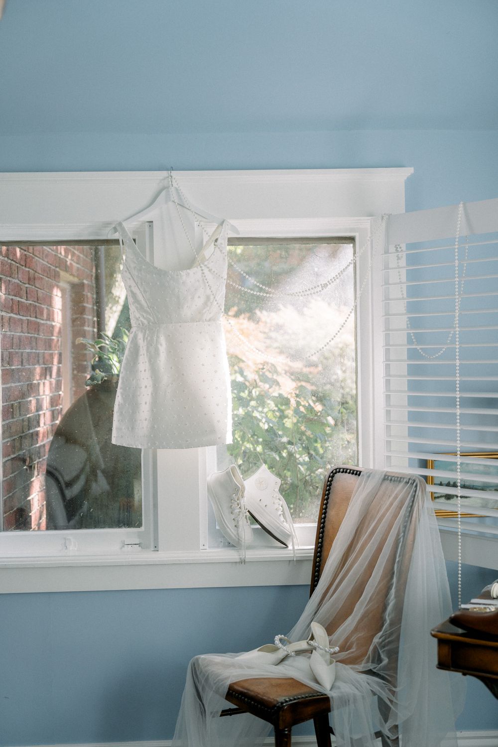 White wedding dress and veil hanging by a window in a blue-walled room with vintage wooden chair and soft natural light.