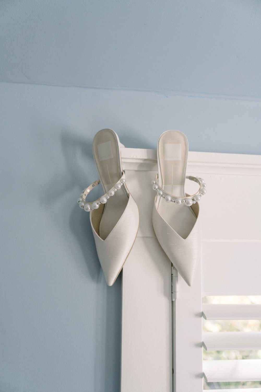 White pearl-adorned bridal heels perched elegantly on a door frame against a pale blue wall.