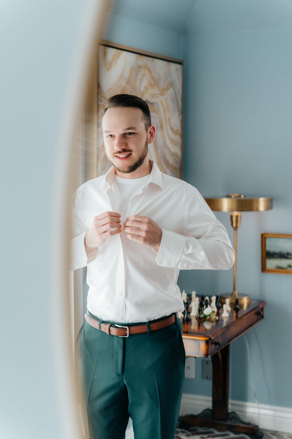 A well-dressed person in a white button-down shirt and green trousers buttons their cuffs in a room with modern decor.